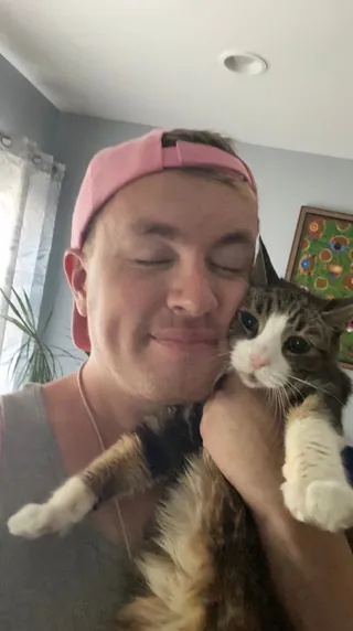 Edward in a pink backwards cap, eyes closed, holding Angel, a tabby and white cat, against his cheek