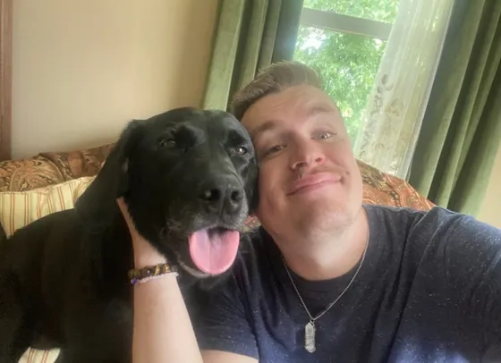 Edward cheek to cheek with Jackie, a black Labrador with her tongue out, on a sofa by a sunlit window