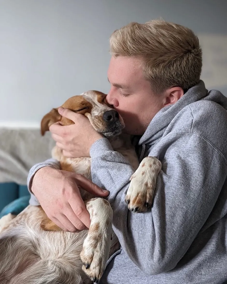 Edward in a grey hoodie, eyes closed, kissing the head of a tan-and-white speckled hound curled against his chest