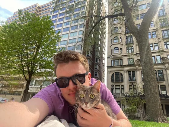 Edward in sunglasses holding Stellaluna, a grey tabby cat, on the grass at Rittenhouse Square with city buildings behind them